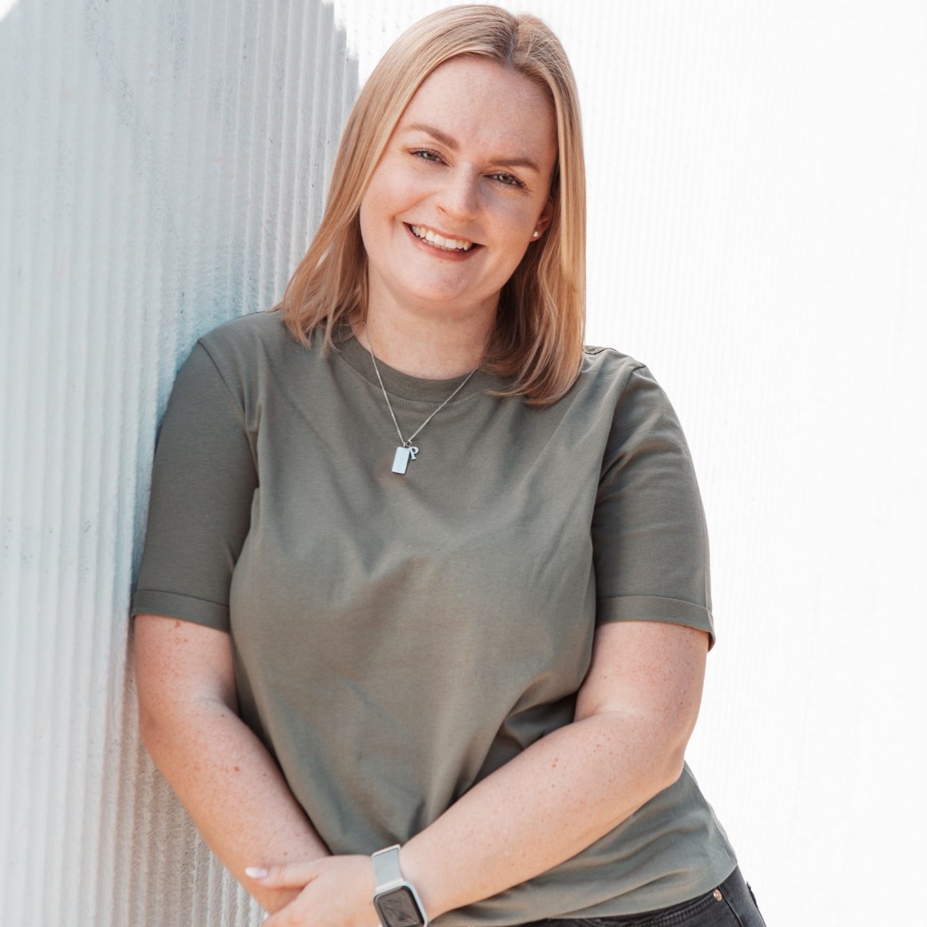 Junge Frau mit blonden Haaren lächelt, trägt ein grünes Shirt und steht lässig an einer Wand.