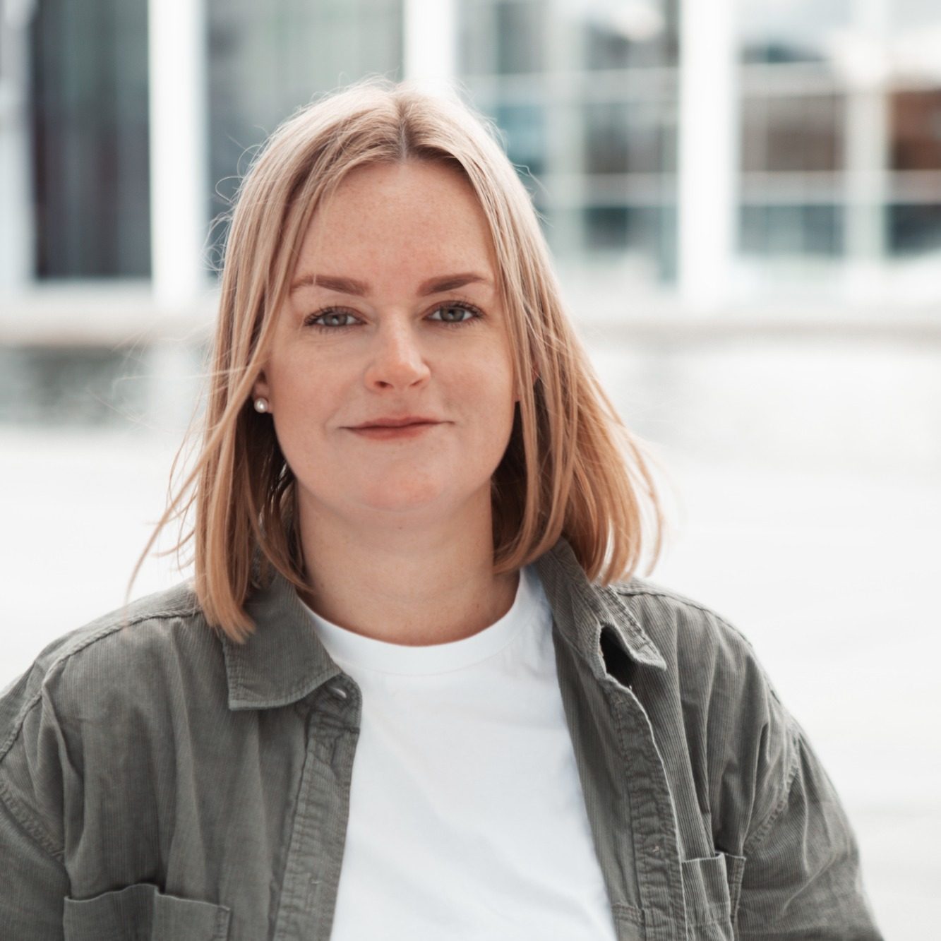 Junge Frau mit blonden Haaren und lässiger Kleidung steht vor einer modernen Glasfassade.